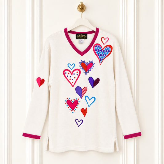 An off-white long-sleeve V-neck tunic top with a colorful heart pattern and magenta trim, hanging on a wooden hanger against a white paneled door.