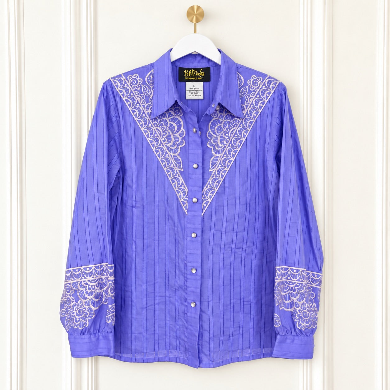 A purple long-sleeve button-up shirt with white embroidered yoke and cuff details, hanging on a white hanger with a gold hook against a cream paneled door.