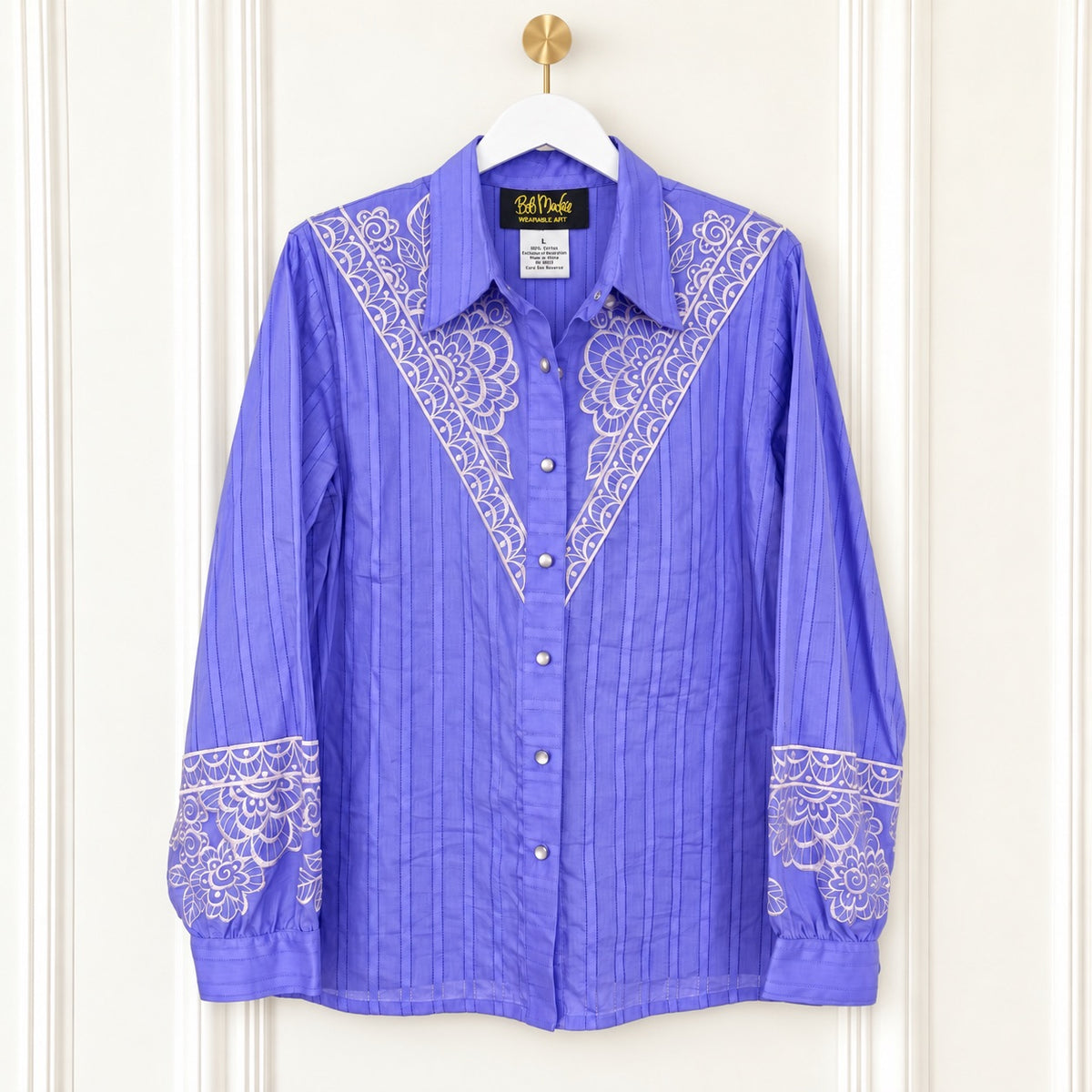 A purple long-sleeve button-up shirt with white embroidered yoke and cuff details, hanging on a white hanger with a gold hook against a cream paneled door.