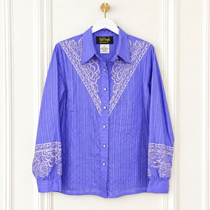 A purple long-sleeve button-up shirt with white embroidered yoke and cuff details, hanging on a white hanger with a gold hook against a cream paneled door.