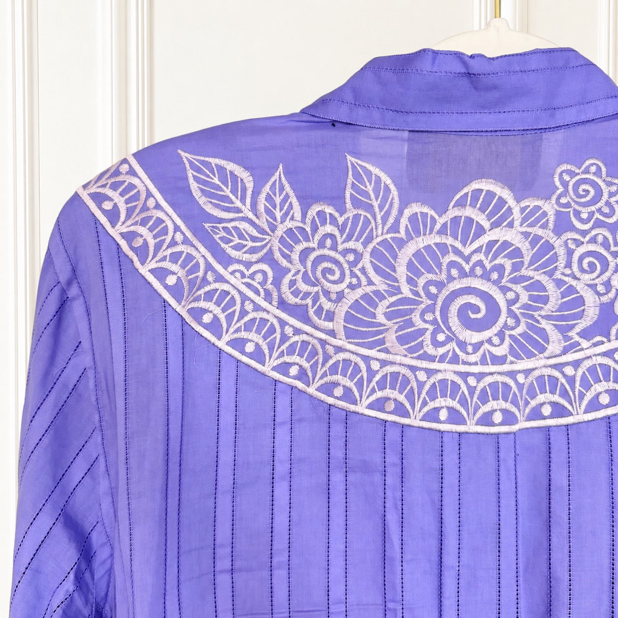 A close-up of a lavender purple blouse with white floral embroidery across the yoke and vertical pleated detailing on the sleeves, shown hanging against a white paneled background.