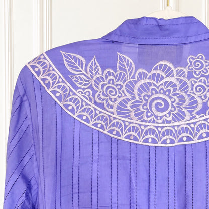 A close-up of a lavender purple blouse with white floral embroidery across the yoke and vertical pleated detailing on the sleeves, shown hanging against a white paneled background.