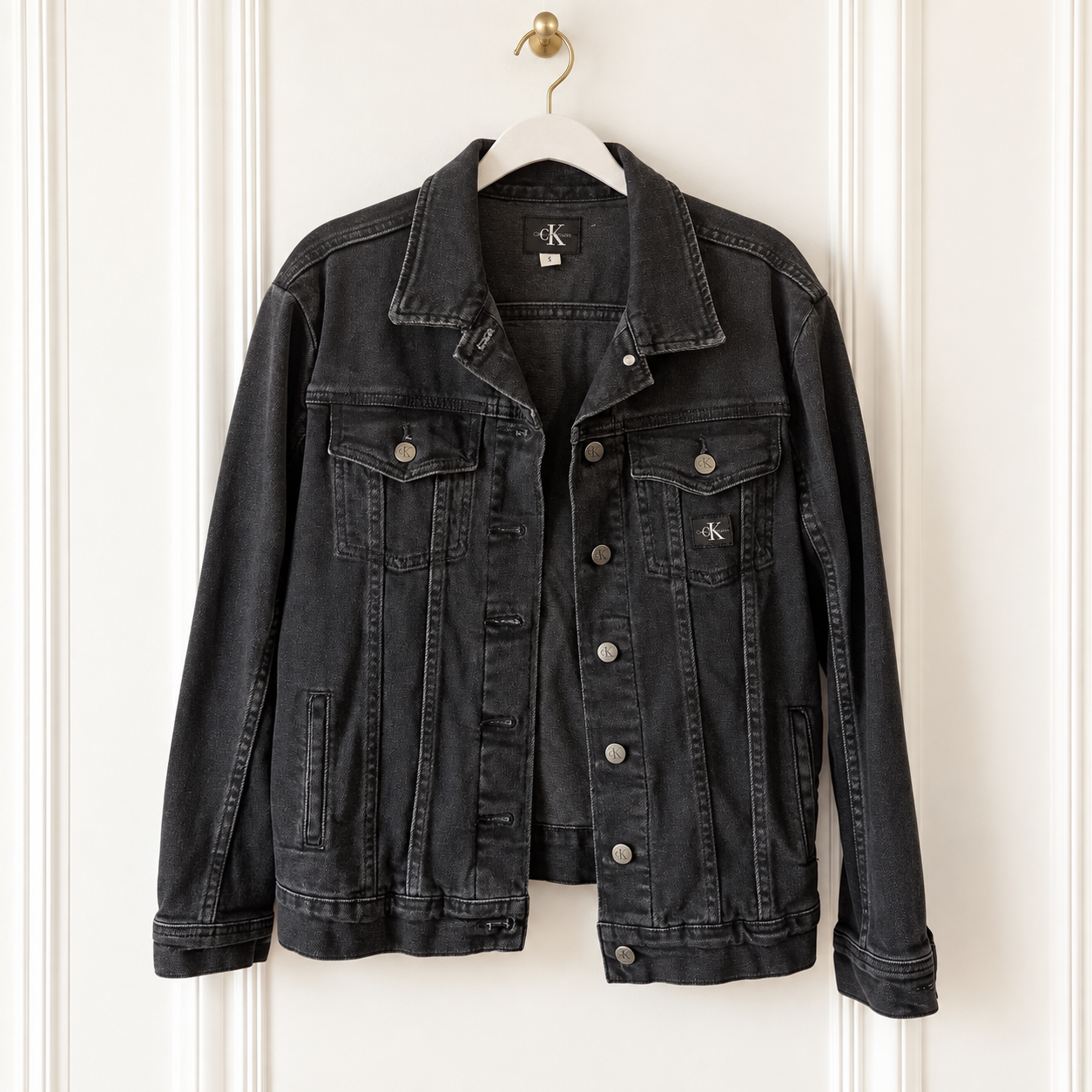 A washed black denim jacket from Calvin Klein hanging on a white hanger against a white paneled door. The jacket features a classic collar, button-down front, and silver-toned metal buttons.
