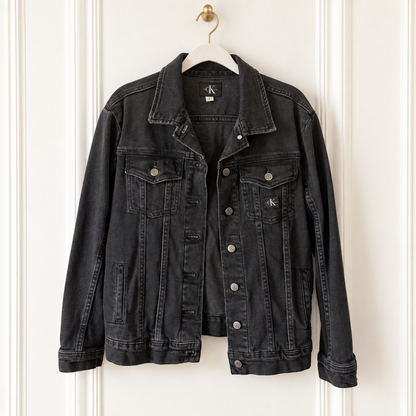 A washed black denim jacket from Calvin Klein hanging on a white hanger against a white paneled door. The jacket features a classic collar, button-down front, and silver-toned metal buttons.