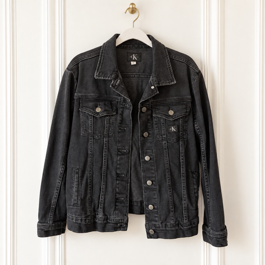 A washed black denim jacket from Calvin Klein hanging on a white hanger against a white paneled door. The jacket features a classic collar, button-down front, and silver-toned metal buttons.