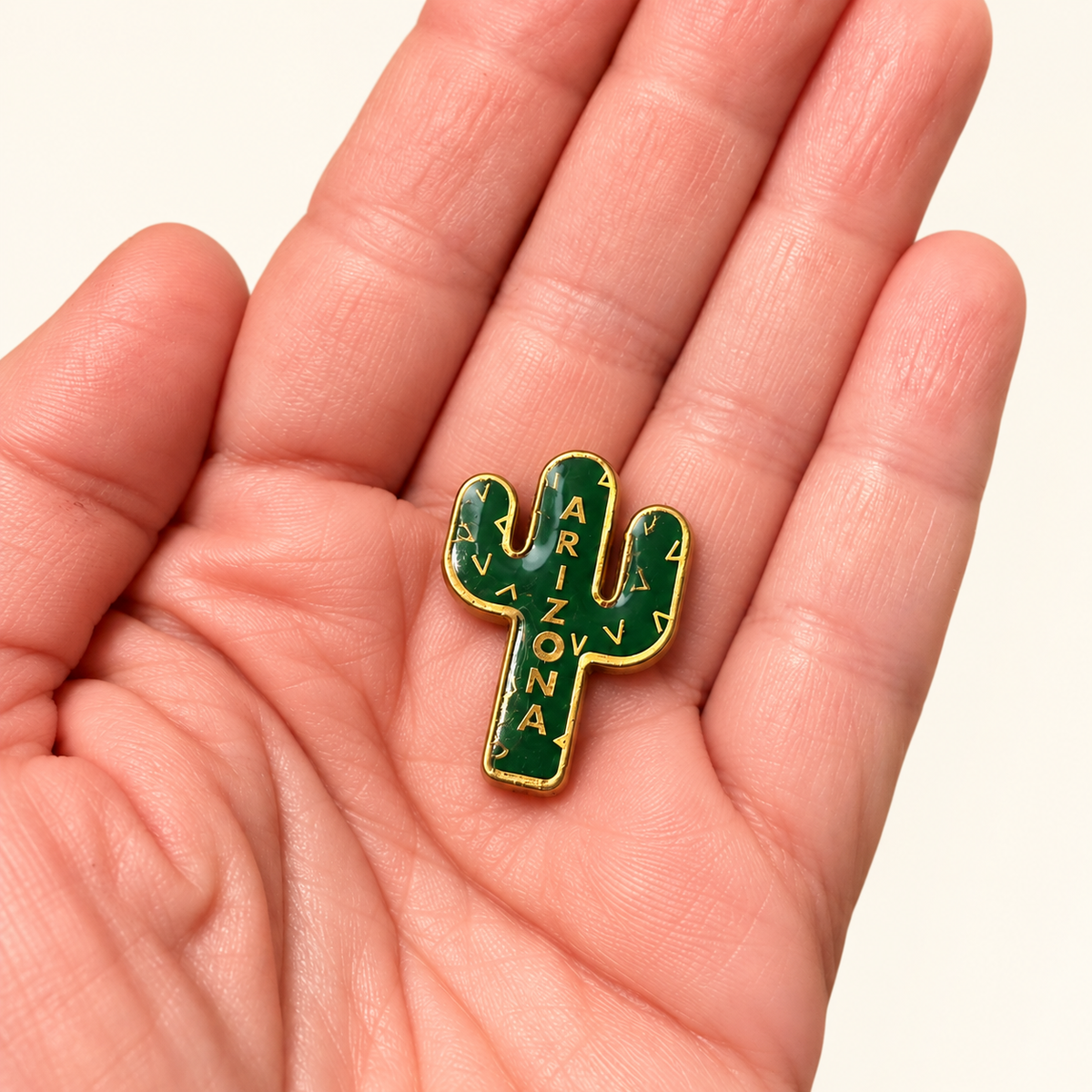 A small cactus-shaped enamel pin with green enamel, gold outline, and 'ARIZONA' written vertically, resting on an open hand.