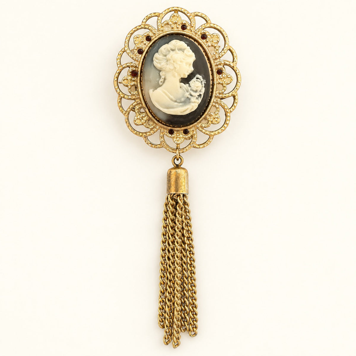 An antique-style gold brooch featuring an oval carved cameo portrait of a woman in profile, surrounded by an ornate filigree frame with small dark gem accents, and a long dangling gold tassel below.