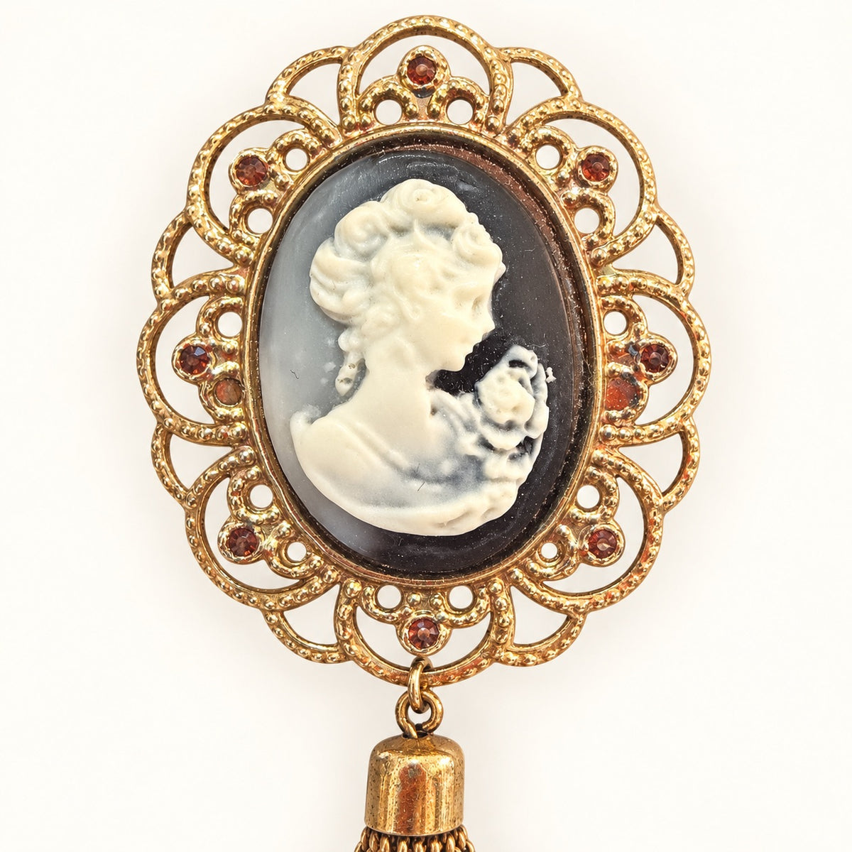 An antique Victorian mourning brooch featuring a carved white cameo profile of a woman with a rose, set in a gold filigree frame with small red gemstone accents and a dangling gold tassel.