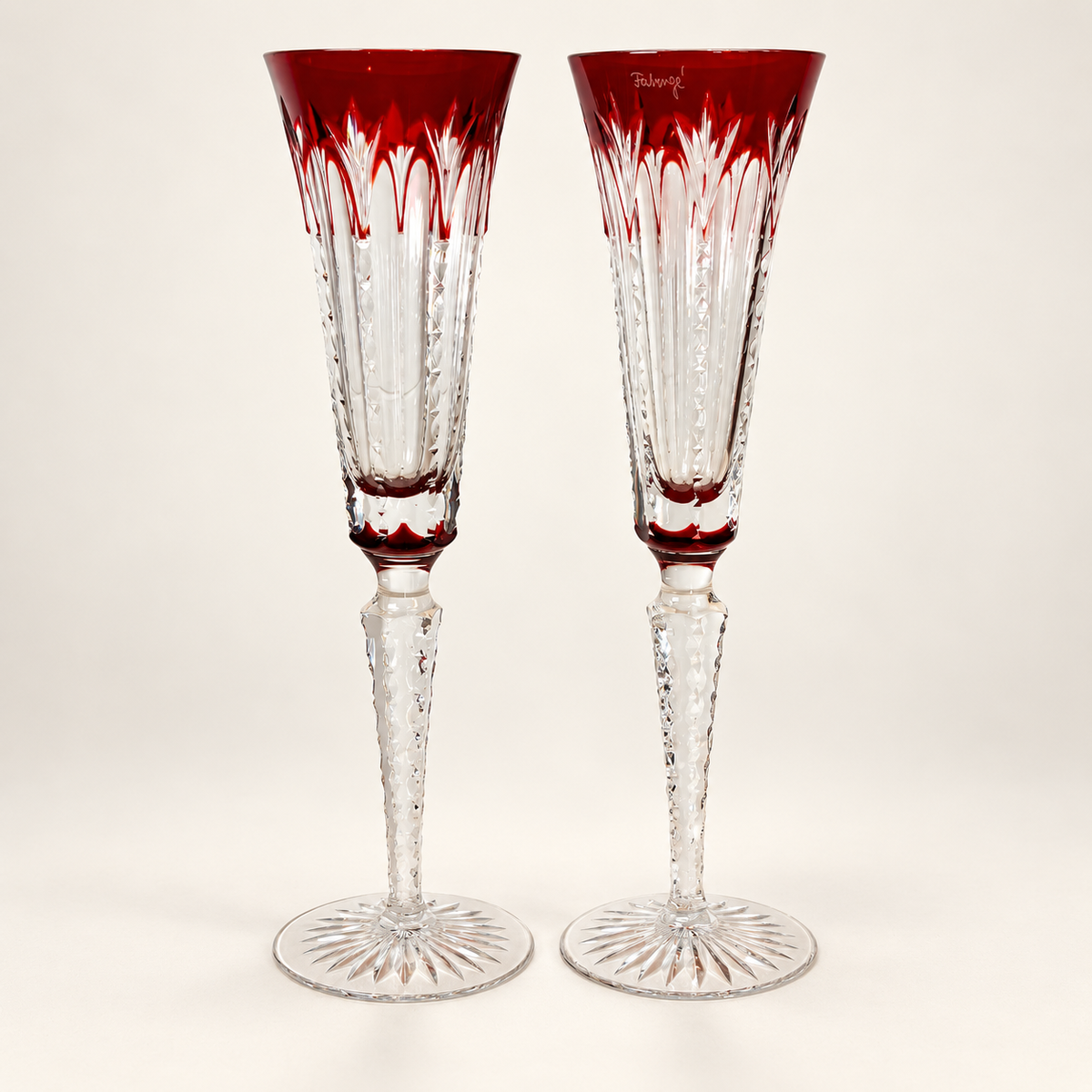 A pair of cut crystal champagne flutes with deep red etched rims and stems, standing on a light beige background.