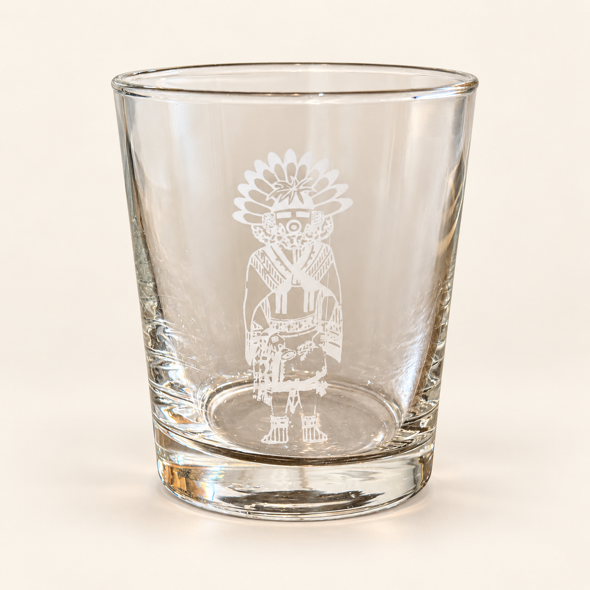 A clear glass rocks tumbler featuring a white etched graphic of a Kachepa figure, shown on a plain off-white background.