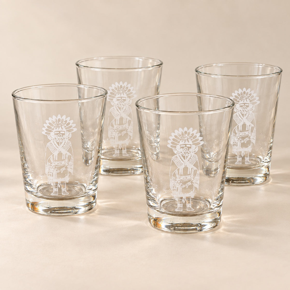 Four clear glass tumblers with white etched Native American-style figures, arranged on a warm beige surface against a matching beige background.