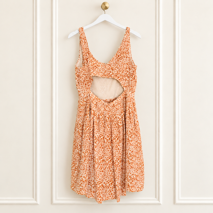 An orange and white floral print sleeveless dress with a cutout at the bust, hanging on a white hanger against a white paneled wall.