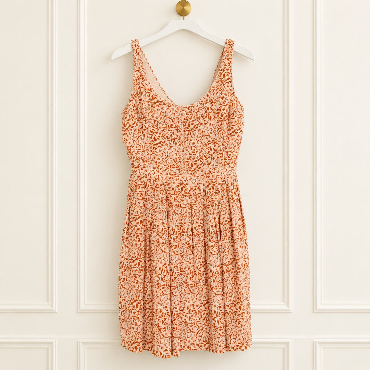 An orange and white floral print sleeveless dress hanging on a white wooden hanger against a white paneled wall.