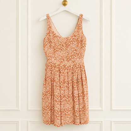 An orange and white floral print sleeveless dress hanging on a white wooden hanger against a white paneled wall.