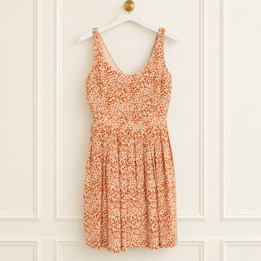 An orange and white floral print sleeveless dress hanging on a white wooden hanger against a white paneled wall.