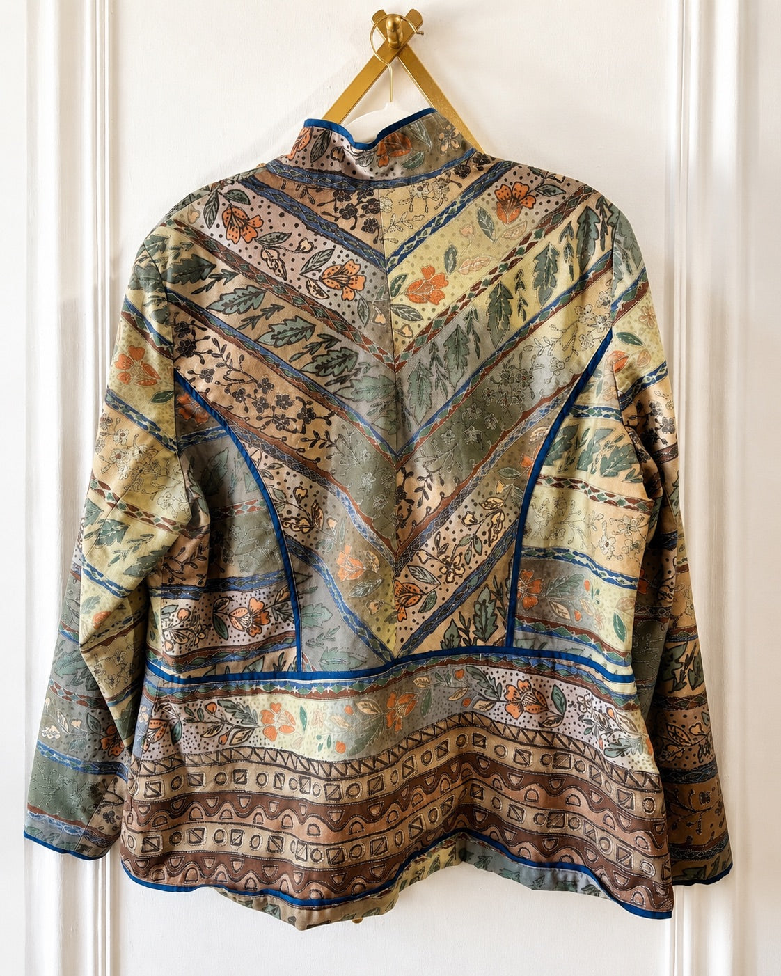 A long-sleeve patterned blouse hanging on a gold hanger against a white paneled wall, showing the back view with a high mock neck and intricate geometric and floral motifs.