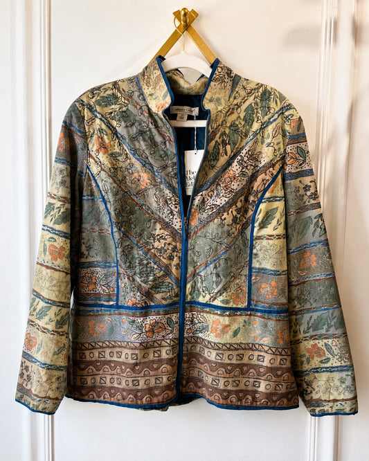 A long-sleeve zip-front jacket with a high stand collar, featuring an intricate multicolor patchwork pattern of blue, green, beige, and brown, hanging on a white hanger against a white paneled door.