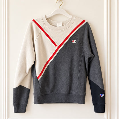 A long-sleeve crewneck sweatshirt hanging on a white hanger against a cream-colored paneled wall. The sweatshirt features a color-blocked design with a dark gray body, a cream upper chest and sleeves, and a diagonal red stripe running from the shoulder to the waist.
