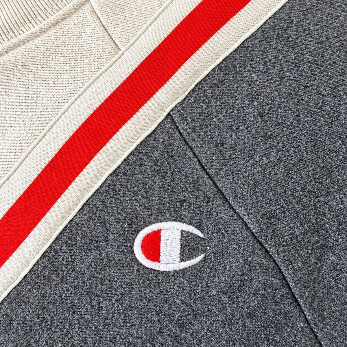 Close-up of a Champion sweatshirt showing a dark heather gray knit body, a cream ribbed collar with a red and cream stripe, and a small embroidered Champion C logo on the chest.