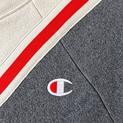 Close-up of a Champion sweatshirt showing a dark heather gray knit body, a cream ribbed collar with a red and cream stripe, and a small embroidered Champion C logo on the chest.