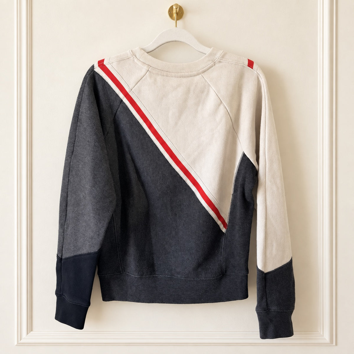 A color-blocked crewneck sweatshirt hanging on a white hanger against a cream-colored paneled wall. The sweatshirt features a diagonal color-blocked pattern with dark gray, off-white, and navy sections, separated by a red and white stripe.
