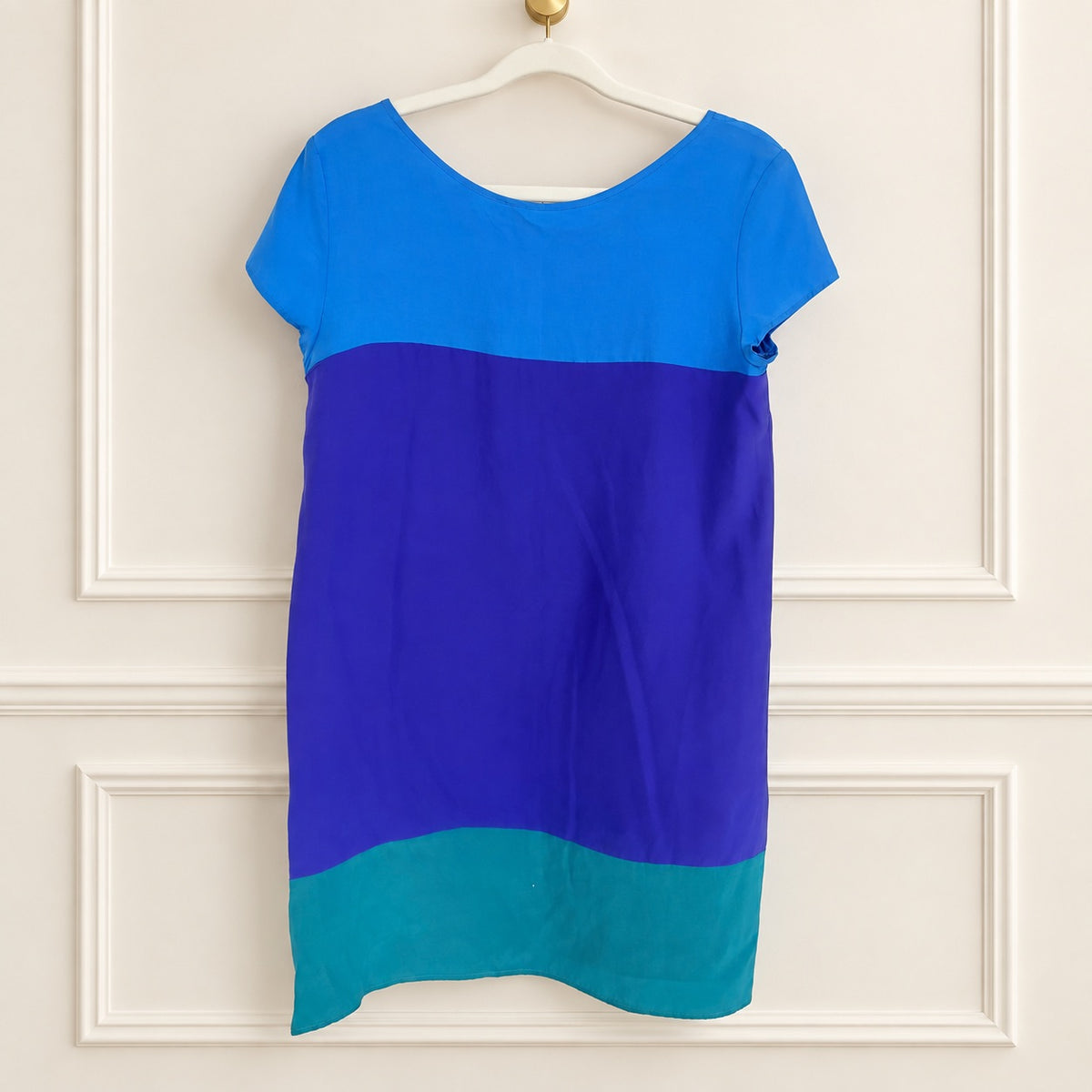A short-sleeve colorblock dress hanging on a white hanger against a white paneled wall. The dress features three horizontal color blocks: a bright blue top section, a large royal purple middle section, and a teal bottom section.