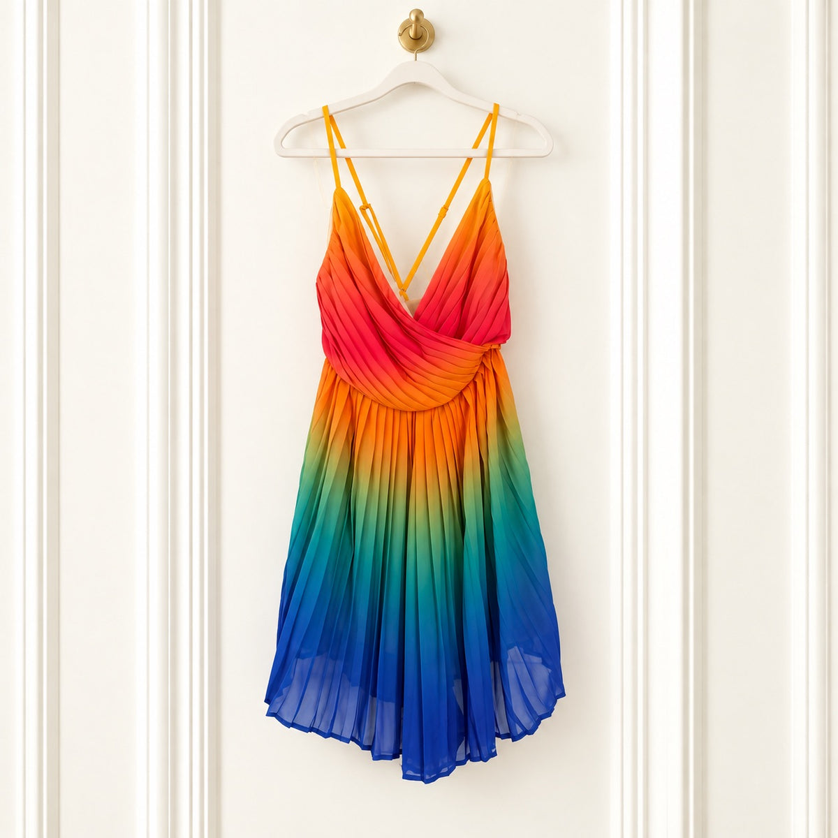 A sleeveless draped dress with thin straps, featuring a vibrant rainbow gradient from red and orange at the top to green, blue, and purple at the bottom, hanging on a white hanger against a white paneled door.