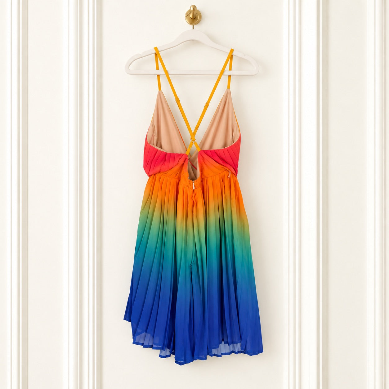 A sleeveless V-neck dress with thin yellow straps, featuring a gradient design that transitions from peach at the top, through orange and pink, to green, blue, and purple at the hem, hanging on a white hanger against a white paneled door.