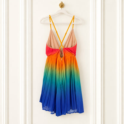A sleeveless V-neck dress with thin yellow straps, featuring a gradient design that transitions from peach at the top, through orange and pink, to green, blue, and purple at the hem, hanging on a white hanger against a white paneled door.