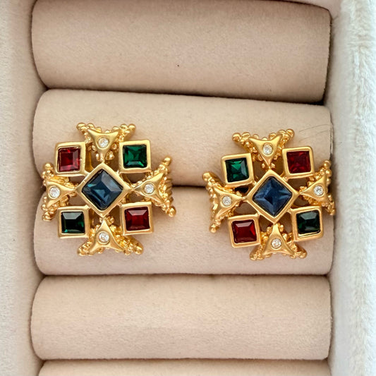 A pair of gold-toned cross-shaped earrings with square red, green, and blue gemstones and small clear accent stones, displayed on a beige jewelry box cushion.