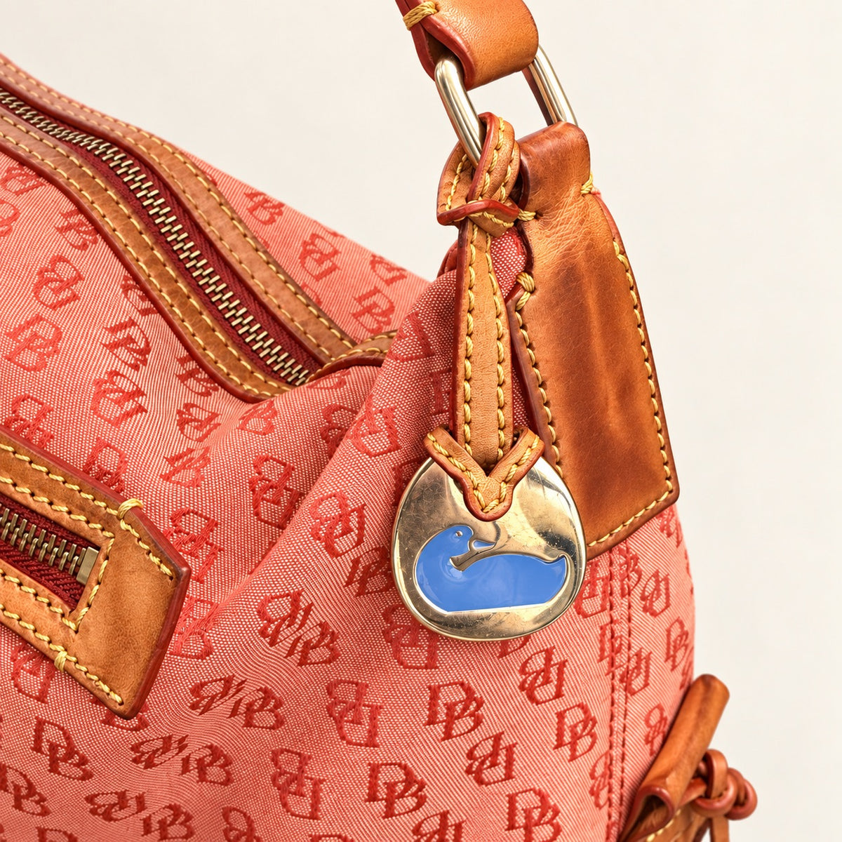 Close-up of a coral pink Dooney bag with an allover red monogram pattern, tan leather trim, and a metal charm featuring a blue duck logo.