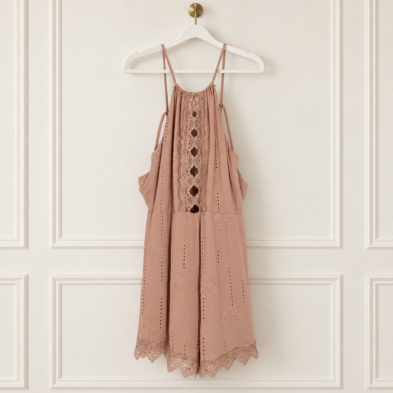 A dusty rose eyelet mini dress with lace trim and button details, hanging on a white hanger against a white paneled wall.