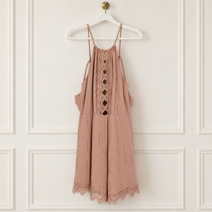A dusty rose eyelet mini dress with lace trim and button details, hanging on a white hanger against a white paneled wall.