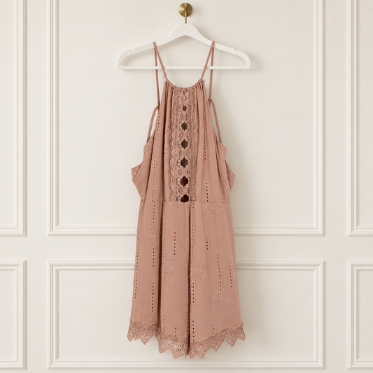 A dusty rose eyelet mini dress with lace trim and button details, hanging on a white hanger against a white paneled wall.
