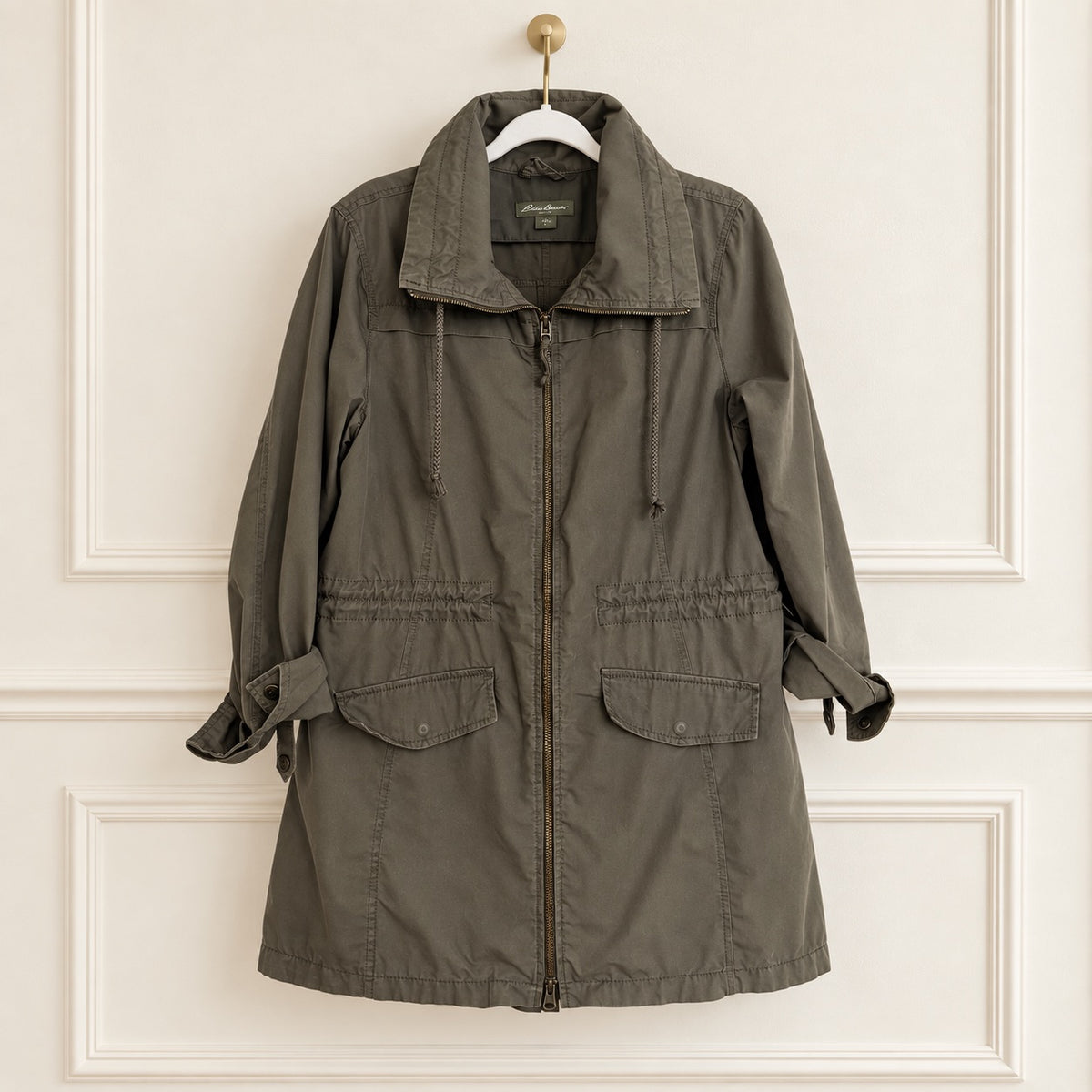 An olive green utility jacket with a high collar, front zipper, and drawstring details, hanging on a white hanger against a cream-colored paneled wall.