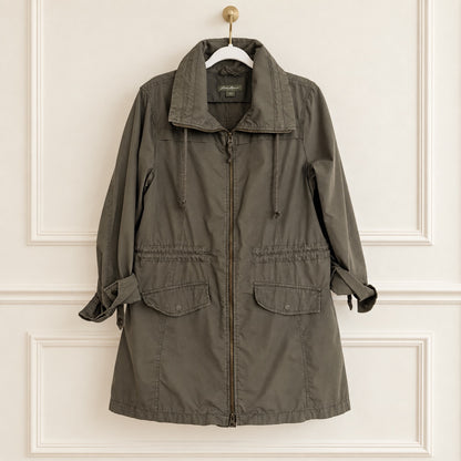 An olive green utility jacket with a high collar, front zipper, and drawstring details, hanging on a white hanger against a cream-colored paneled wall.