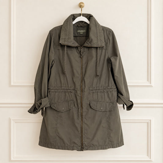 An olive green utility jacket with a high collar, front zipper, and drawstring details, hanging on a white hanger against a cream-colored paneled wall.