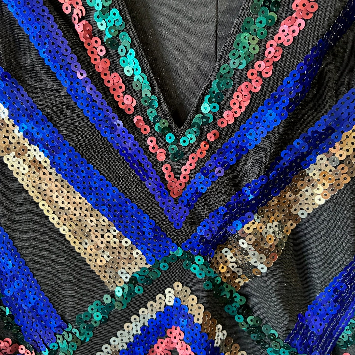 Close-up of a black sequined garment featuring geometric V-shaped and striped patterns of metallic blue, teal green, pink, and gold sequins.