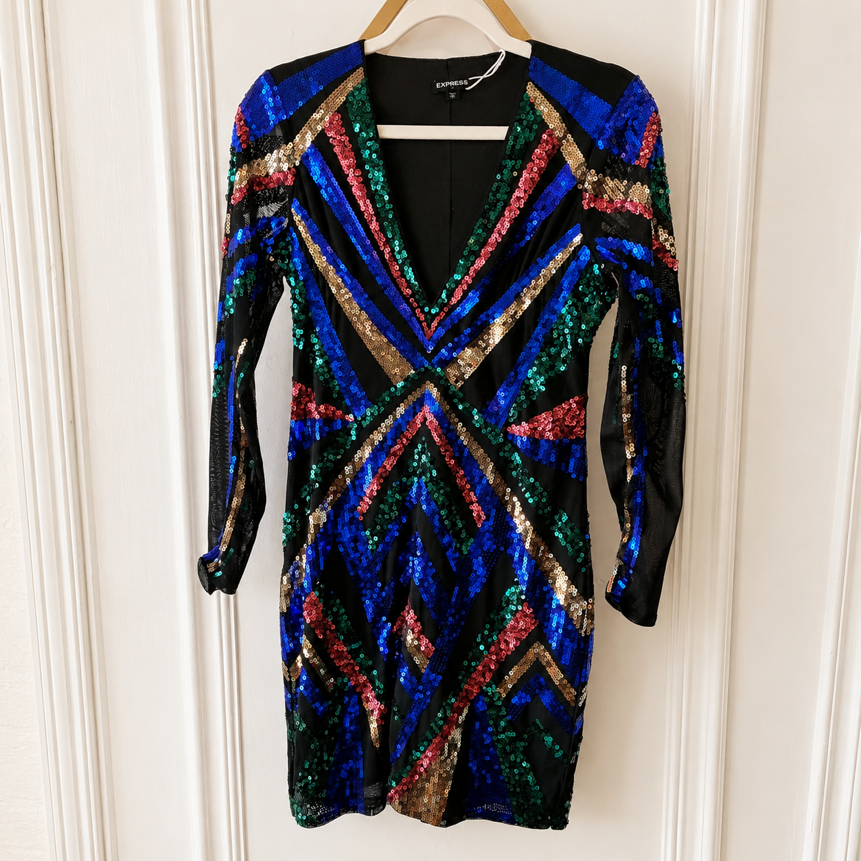 A long-sleeve sequined mini dress hanging on a wooden hanger against a white paneled wall. The dress features a black base covered in vibrant, multicolored sequins in blue, green, red, and gold, arranged in geometric chevron and striped patterns.