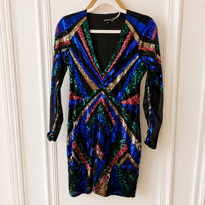 A long-sleeve sequined mini dress hanging on a wooden hanger against a white paneled wall. The dress features a black base covered in vibrant, multicolored sequins in blue, green, red, and gold, arranged in geometric chevron and striped patterns.