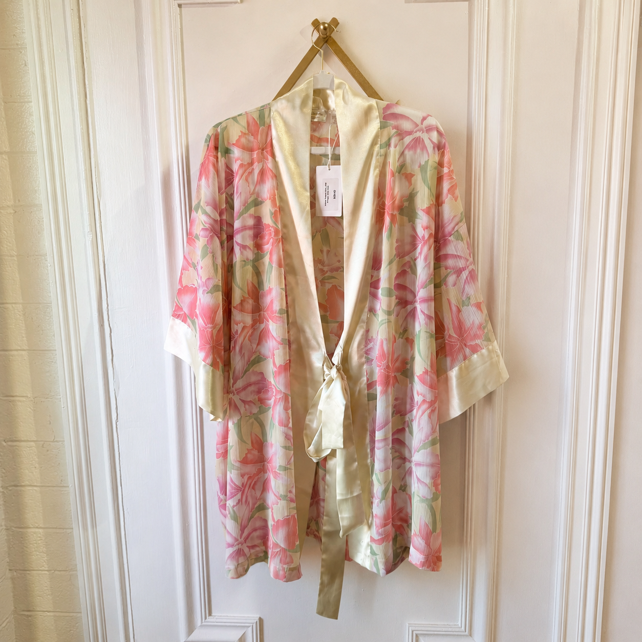 A lightweight floral robe with pink and green floral print and cream satin trim, hanging on a gold hanger against a white paneled door.