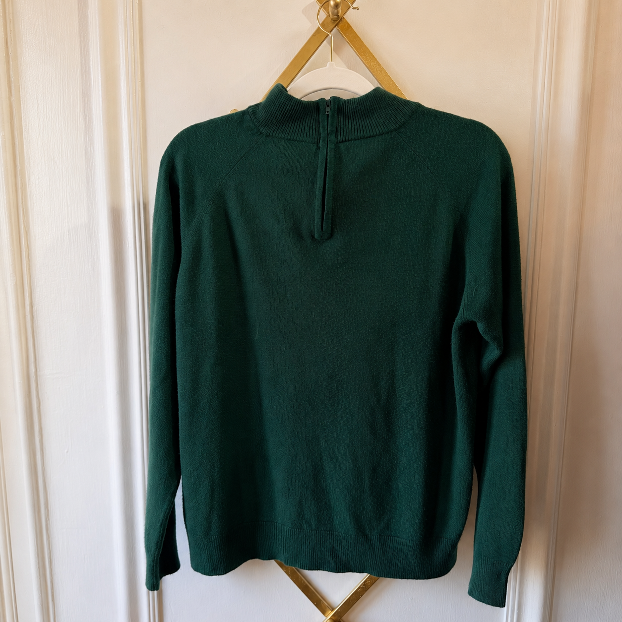 A dark green knit sweater with a mock neck and split neckline, hanging on a gold hanger against a white paneled door.