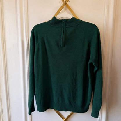 A dark green knit sweater with a mock neck and split neckline, hanging on a gold hanger against a white paneled door.
