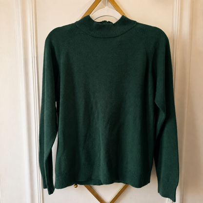 A dark forest green knit sweater with a mock neck and long sleeves, hanging on a white hanger with a gold metal frame against a cream-colored paneled door.
