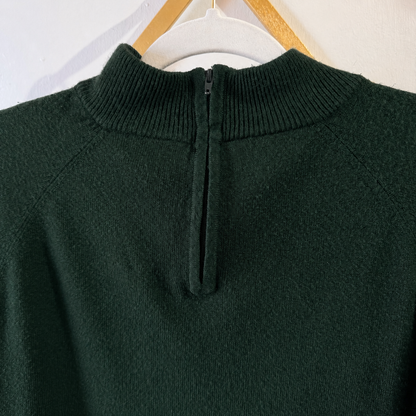 A dark forest green knit sweater with a unique split neckline featuring a central zipper, hanging on a white hanger with a wooden hook against a white wall.
