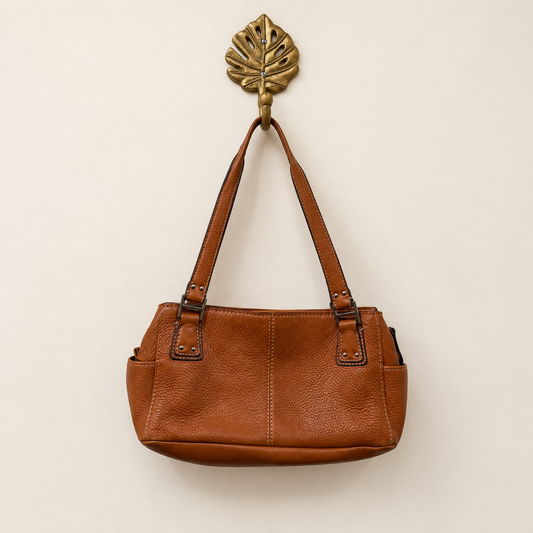 A medium brown pebbled leather shoulder bag hanging from a decorative brass leaf-shaped wall hook against a plain off-white wall.
