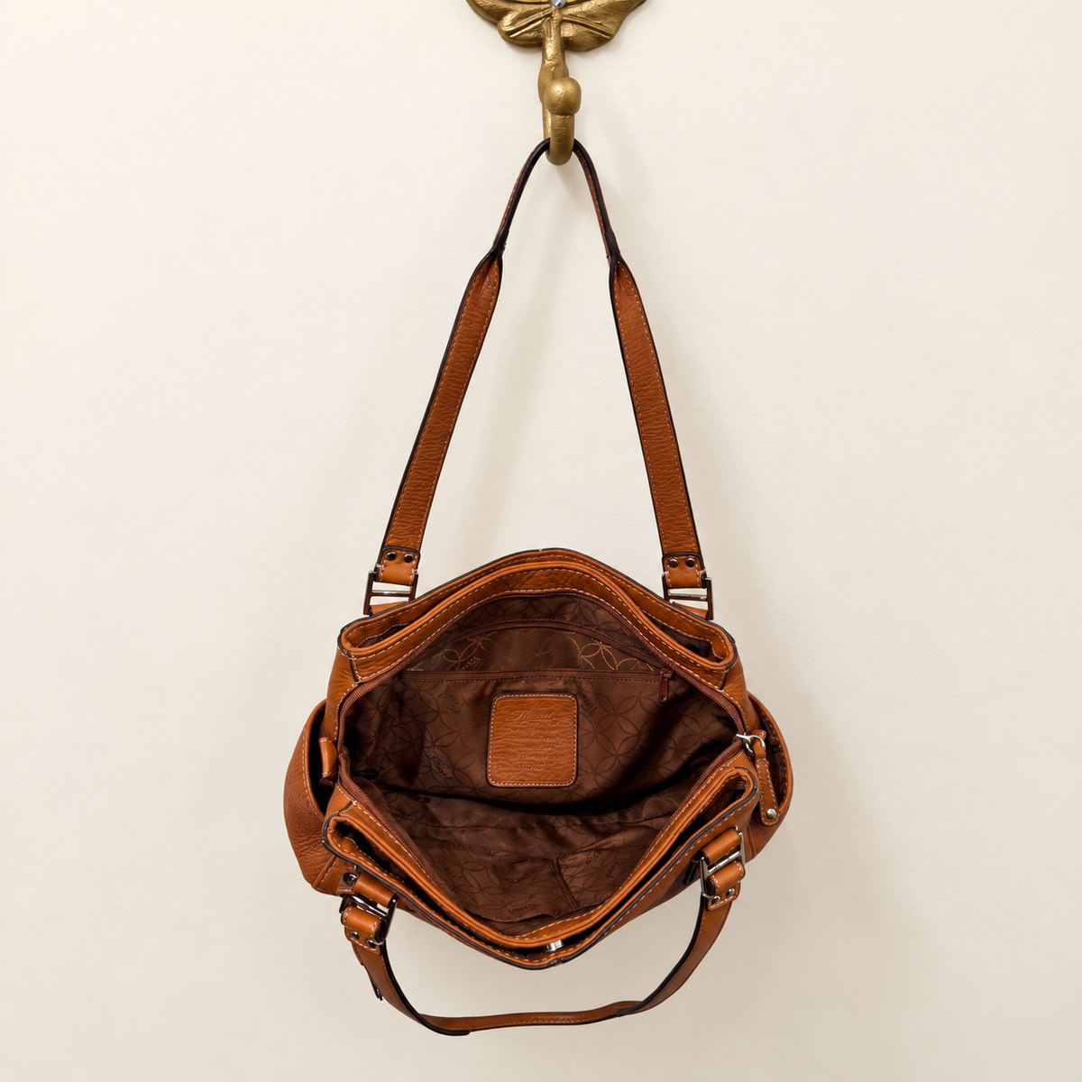 A brown leather shoulder bag with an open top, hanging from a brass wall hook against a plain off-white wall.