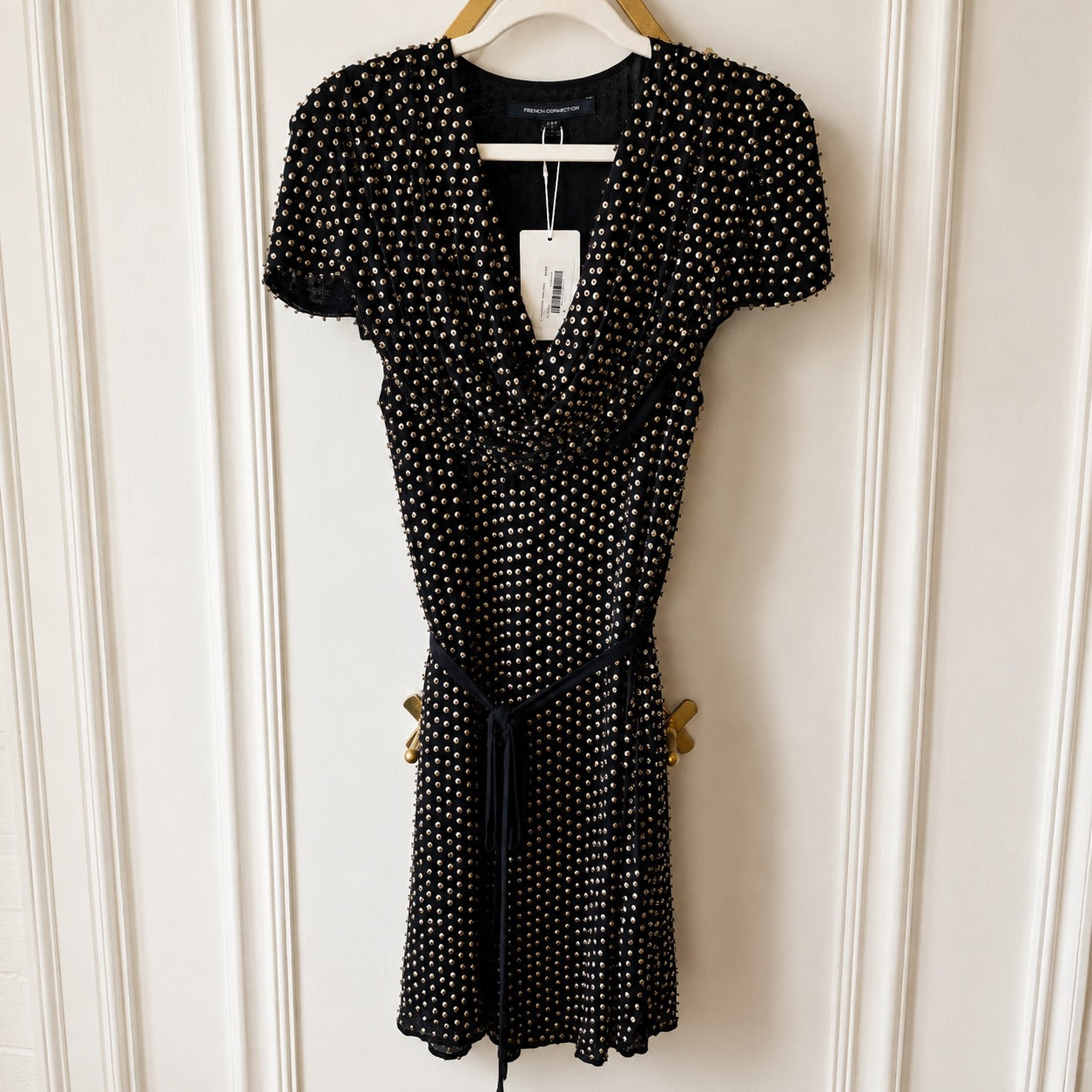 A black short-sleeve wrap-style dress with a white polka dot pattern, hanging on a wooden hanger against a white paneled door. The dress features a tie belt at the waist and a visible H&M logo on the inner neck label.
