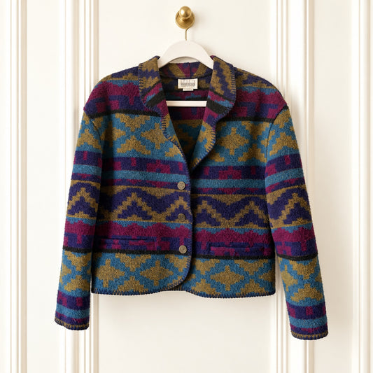 A cropped knit blazer with a colorful geometric pattern in purple, blue, teal, and mustard, hanging on a white hanger against a white paneled door.