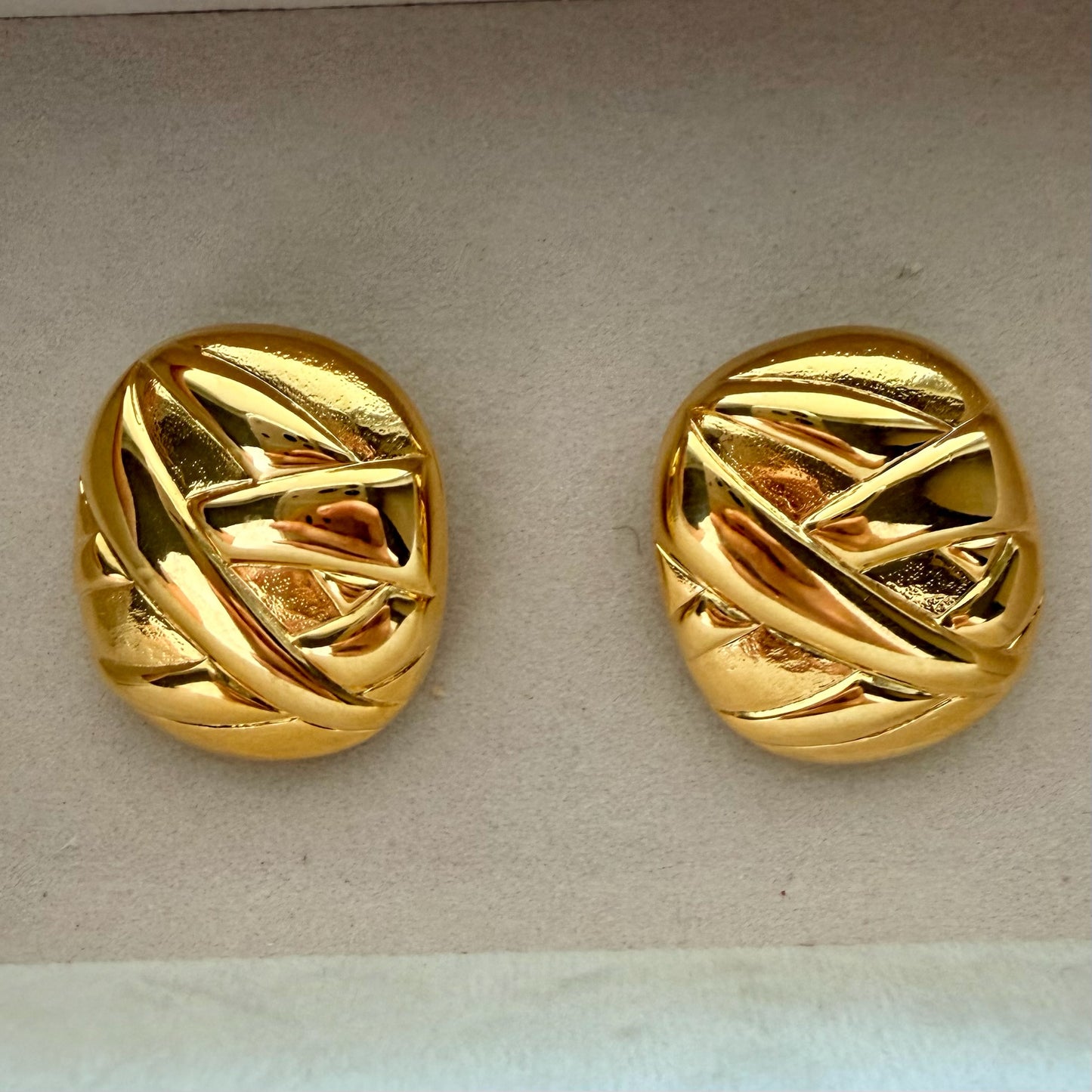 A pair of round gold earrings with an abstract, textured pattern, displayed on a light beige textured surface.